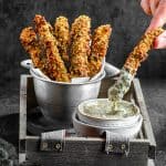 Baked zucchini fries in a fry holder with a small bowl of tahini sauce with a fry being dipped in the sauce