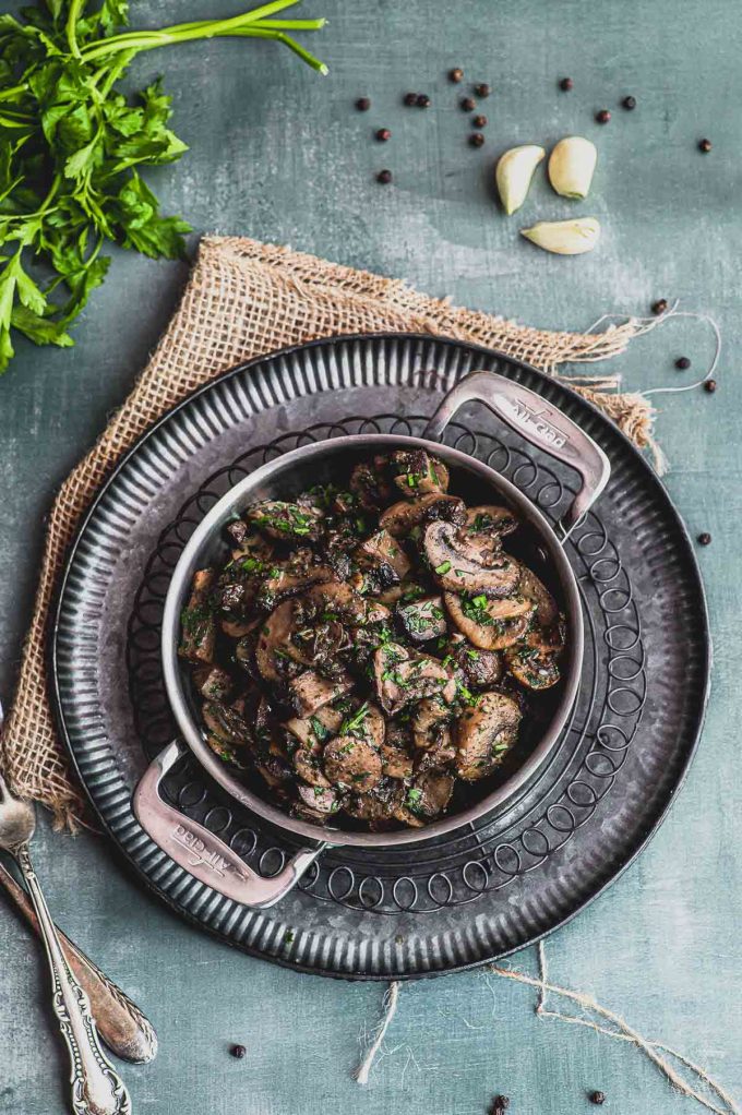 Birds eye view of a mini skillet with sauteed mushrooms next to cloves of garlic and parsley