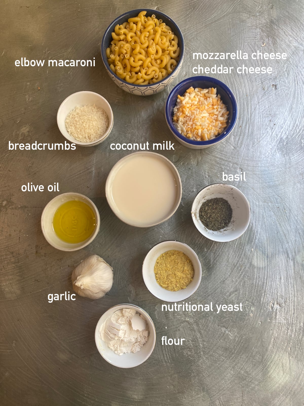 overhead shot of ingredients to make vegan mac and cheese bites. There are 8 small white bowls filled with each ingredient and a whole bulb of garlic set on a grey stone background