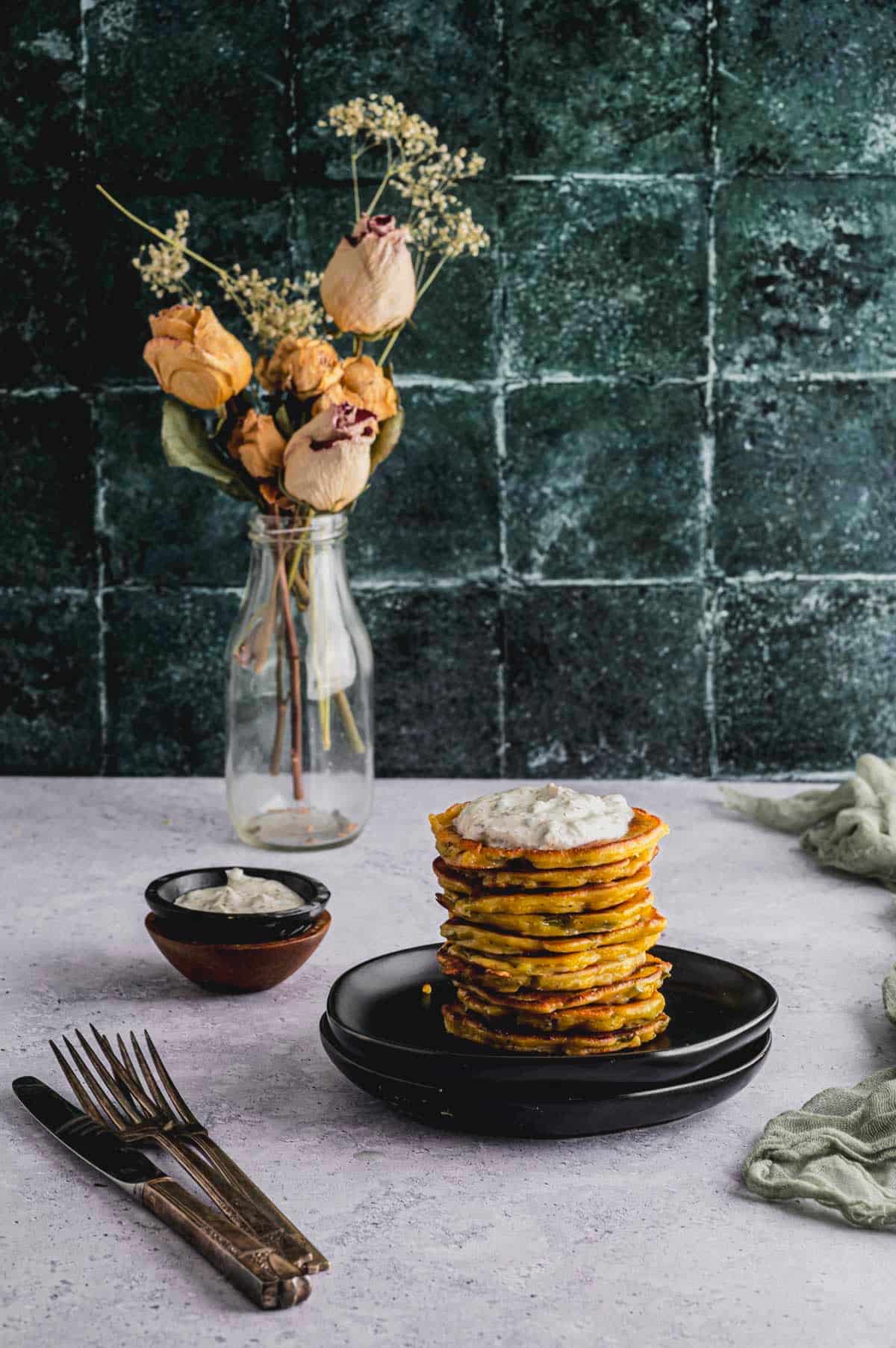 A stack of Savory Chickpea Flour and Scallion Pancakes plated on a small black plate. A small bowl of Greek Yogurt Caper Sauce and a vase of flowers sit in the background.