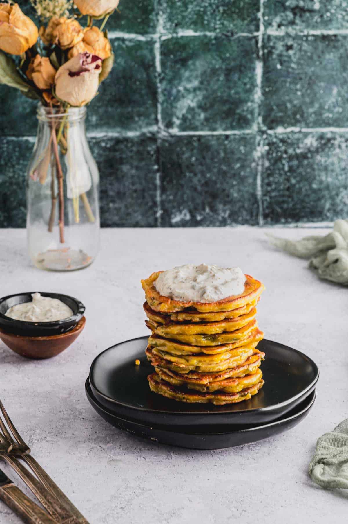 A stack of Savory Chickpea Flour and Scallion Pancakes plated on a small black plate. A small bowl of Greek Yogurt Caper Sauce and a vase of flowers sit in the background.