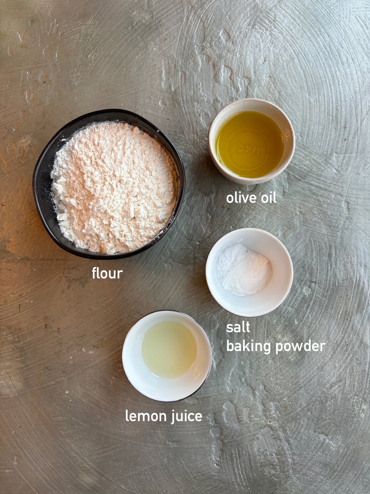 overhead shot of the ingredients needed to make the dough for Fatayer Spinach Pies. Each ingredient is in a small bowl and labeled with white text. 
