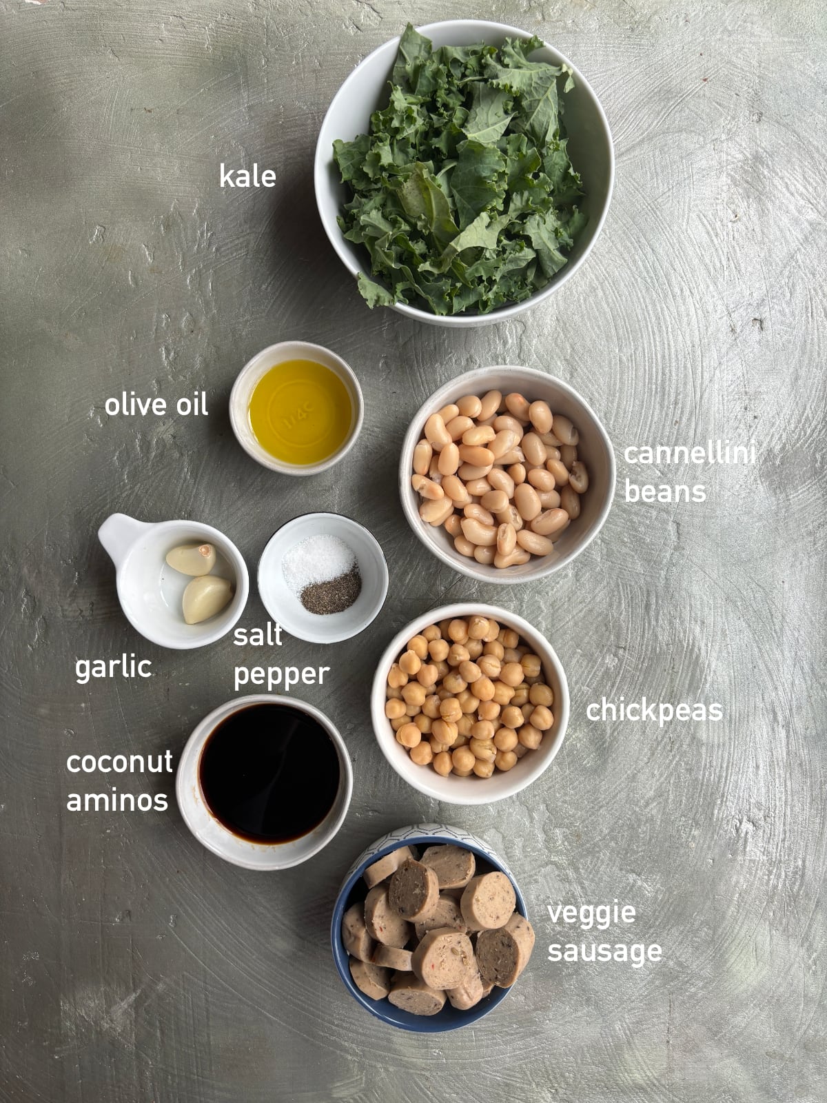 overhead shot of several small bowls filled with the ingredients needed to make kale soup with veggie sausage. The bowls are labeled with the name of each ingredient. 