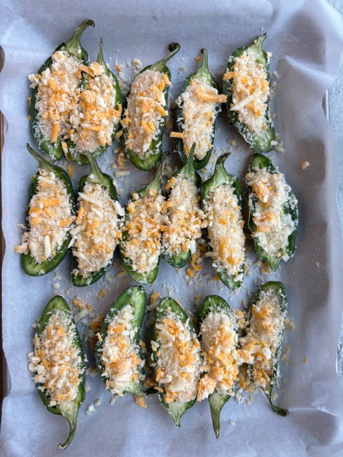 overhead shot of jalapeño halves stuffed with cheese and topped with breadcrumbs on a parchment paper lined sheet tray before being baked