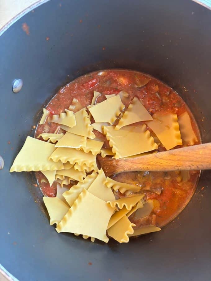 overhead image of nonstick pot cooking chunky tomato soup and broken lasagna noodles with a wooden spoon