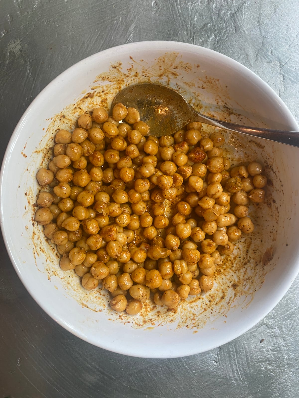 overhead view of a white mixing bowl with chickpeas being tossed with oil and spices with a metal spoon