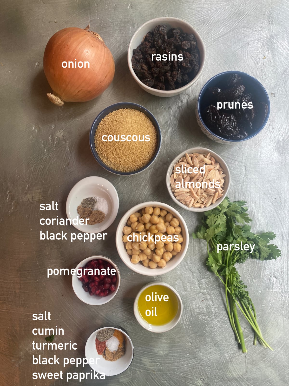 overhead image of the ingredients needed to make moroccan couscous. Each ingredient is in it's own small bowl and labeled with white text