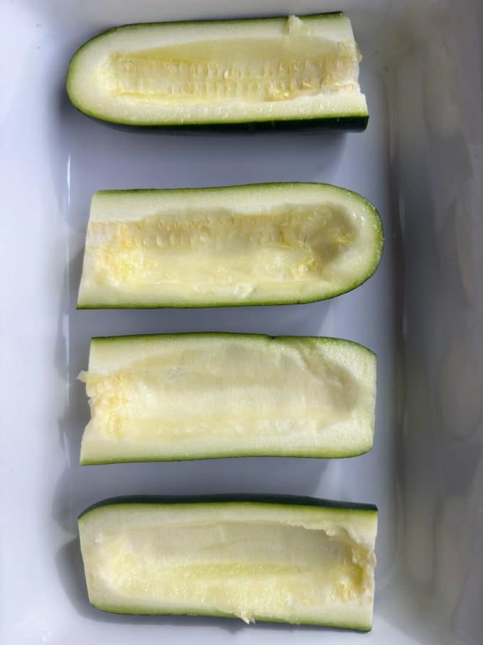 overhead image of 4 zucchini halves sliced lengthwise and the middles scooped out to form boat shapes. They are in a white baking dish.