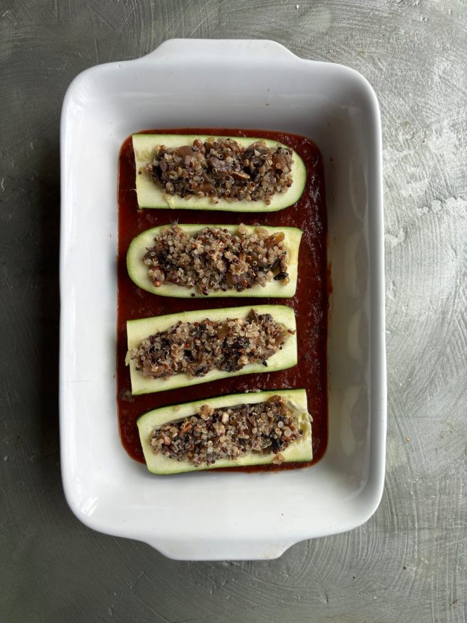 overhead image of a white baking dish with a layer of tomato sauce inside. Zucchini boats filled with quinoa and vegetable filling.