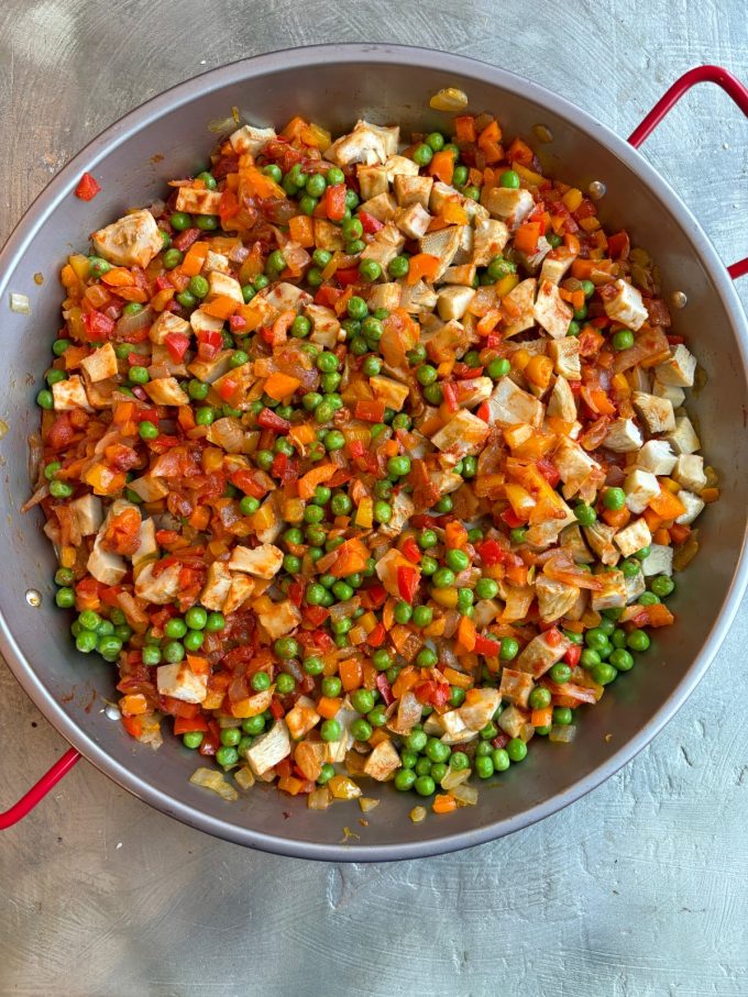 overhead image of a paella pan filled with a mixture of diced red, orange and yellow bell peppers, diced onions, green peas, diced artichoke bottoms.