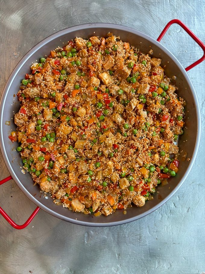 overhead image of a paella pan filled with a mixture of diced red, orange and yellow bell peppers, minced garlic, diced onions, green peas, diced artichoke bottoms, quinoa