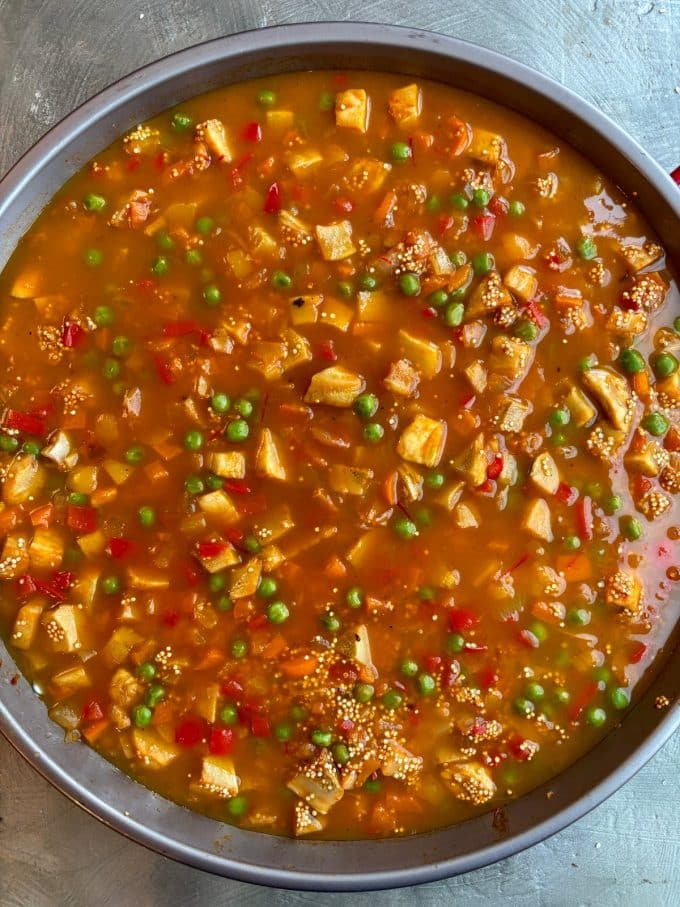 overhead image of a paella pan filled with a mixture of diced red, orange and yellow bell peppers, diced onions, green peas, diced artichoke bottoms, quinoa, vegetable broth
