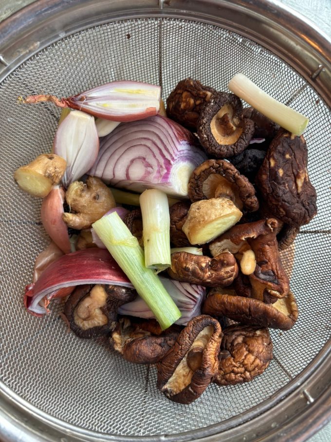 overhead image of mushrooms, onions, scallions, and ginger in a fine mesh sieve