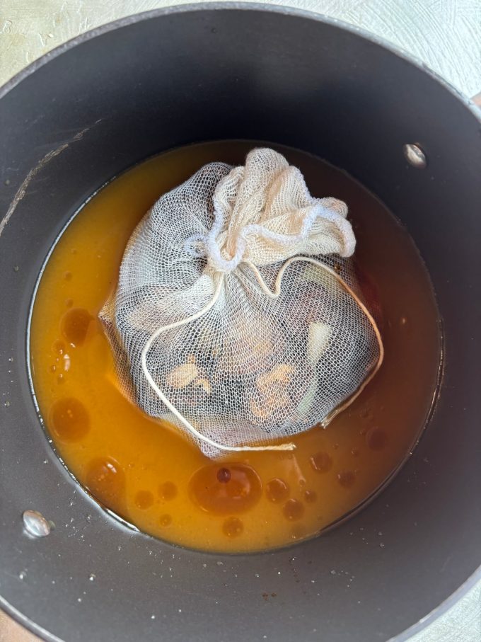 overhead image of chopped mushrooms, onions, scallions and ginger wrapped in cheesecloth sitting in an orange-hued broth inside a nonstick pot