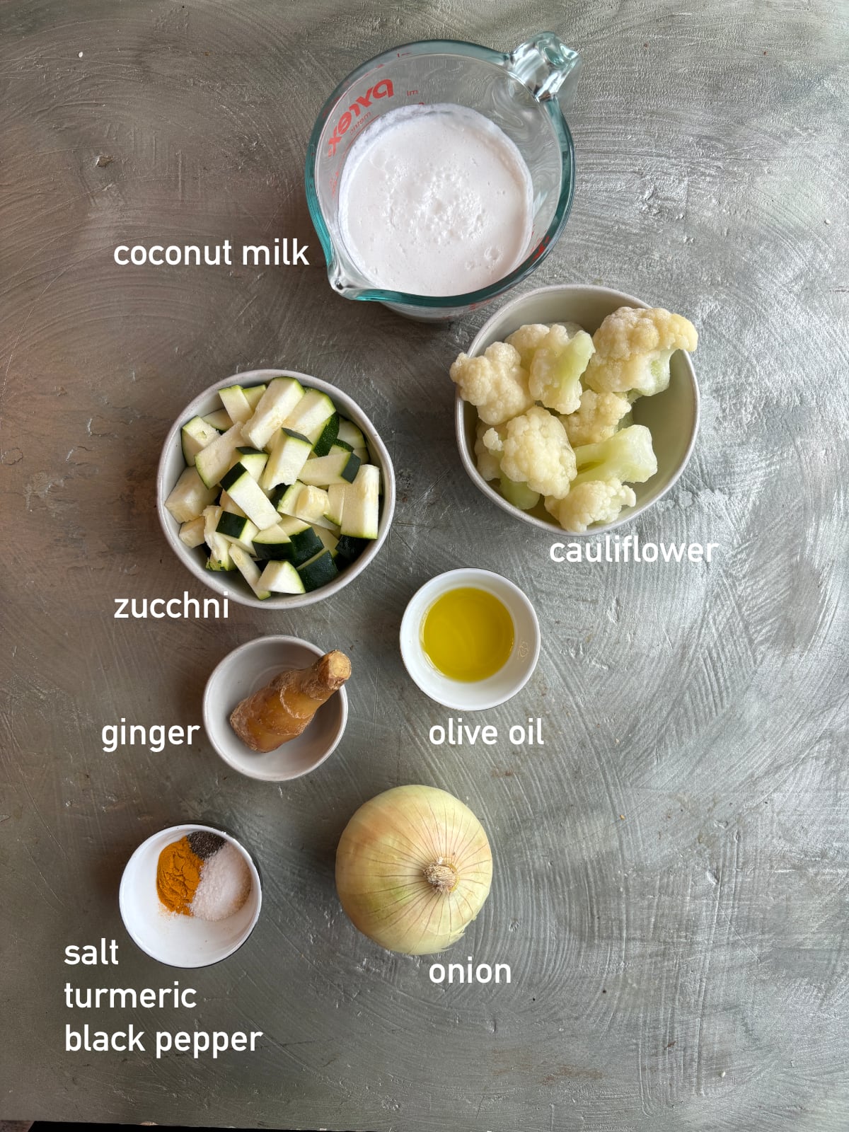 overhead image of the ingredients needed to make zucchini cauliflower soup. Each ingredient is in its own bowl and labeled with white text
