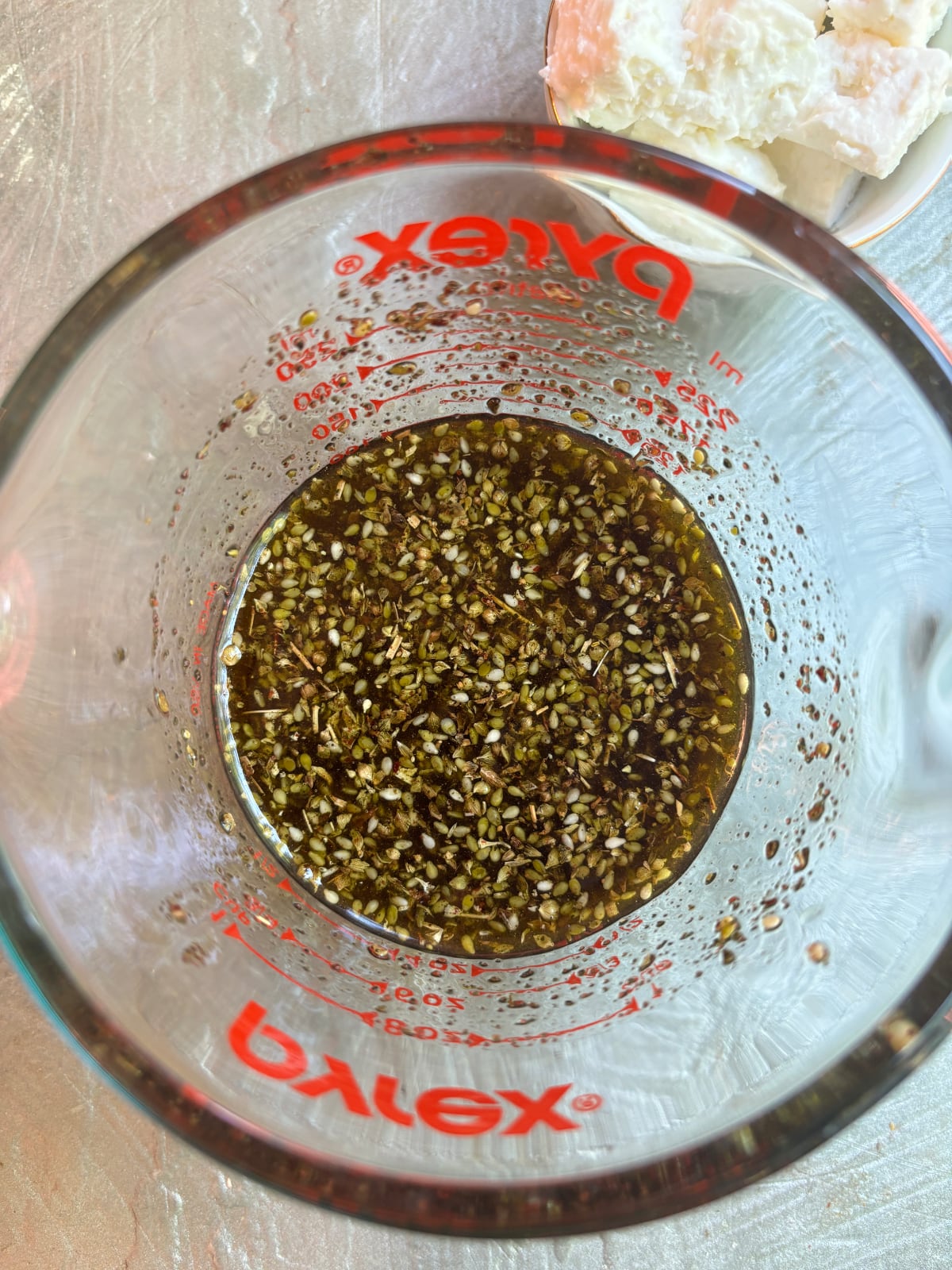 overhead image of a glass liquid measuring cup. Inside is a mixture of oil and herbs for Marinated Feta Cheese.