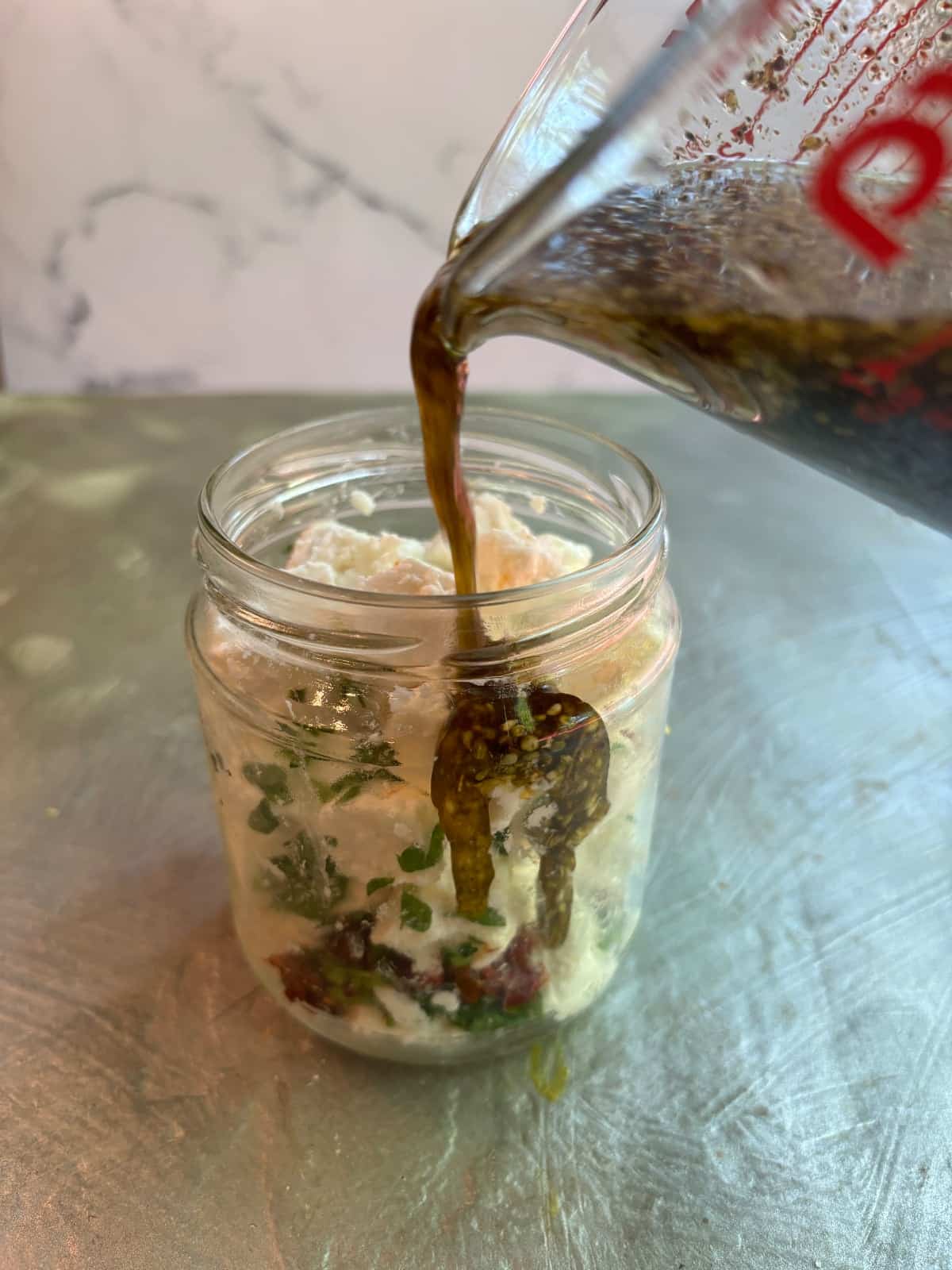 Cubed feta cheese in a glass jar with the marinade being poured over it