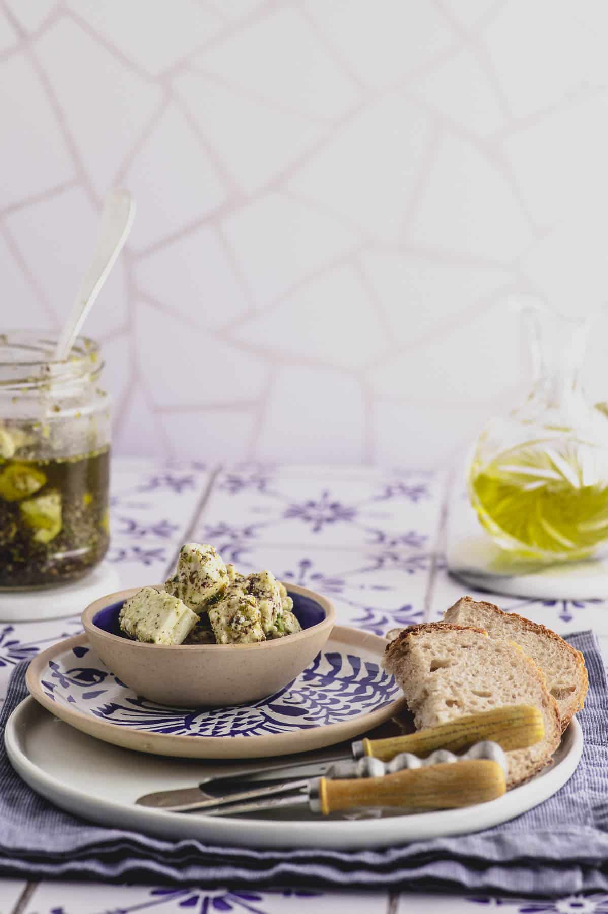 a plate with bread and a small bowl with marinated feta cheese a bottle of olive oil and a jar with feta in the background
