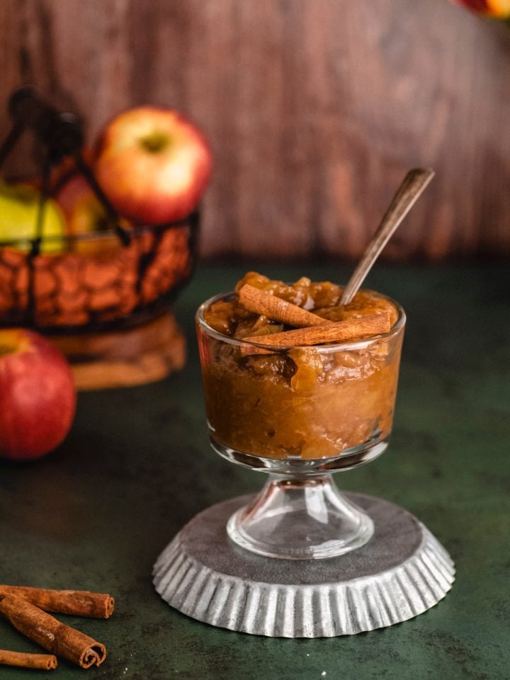 a glass cup holding dark golden brown apple butter, topped with whole cinnamon sticks. There is a basket of apples in the background.