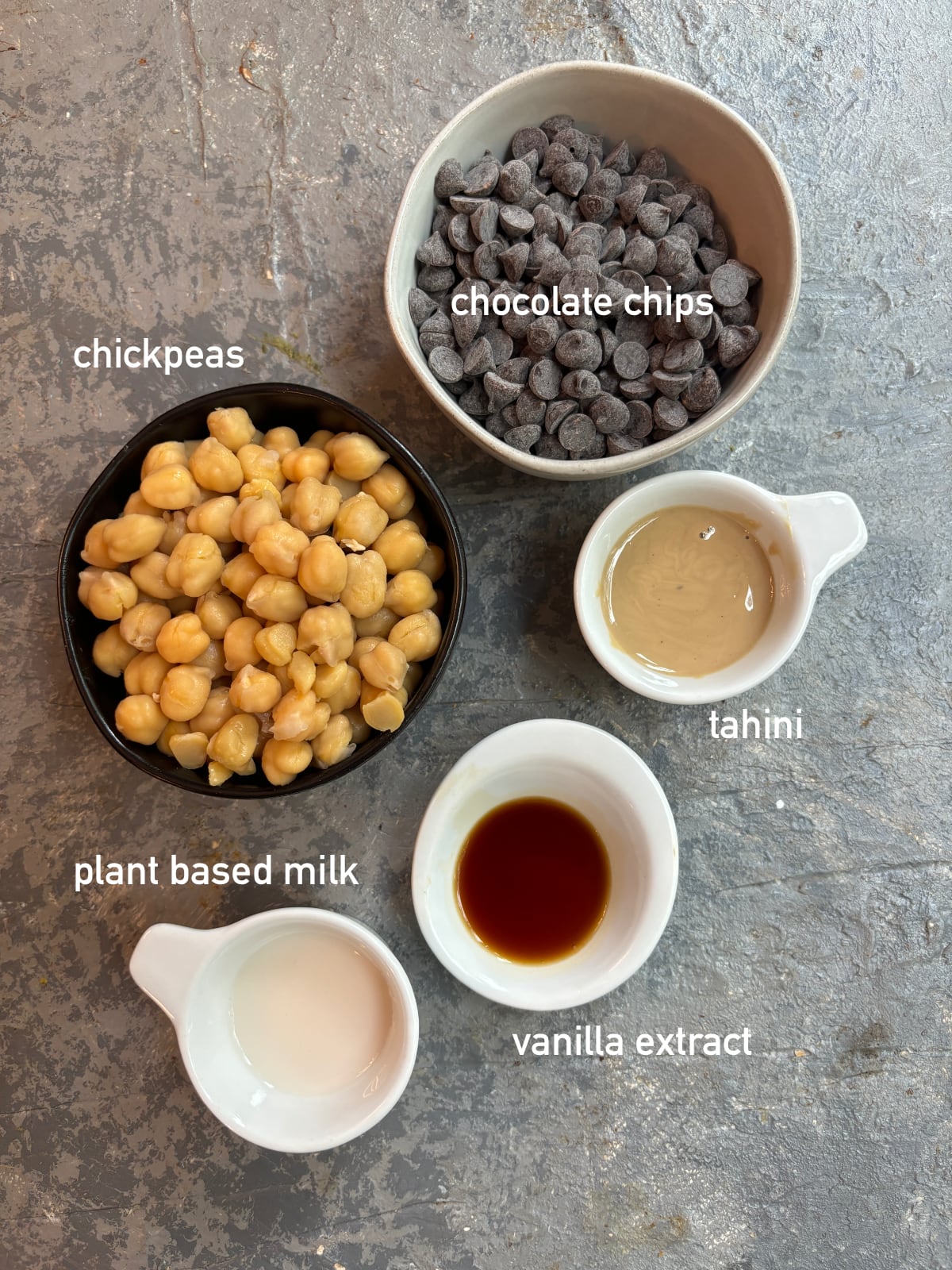overhead image of the ingredients needed to make chocolate hummus. Each ingredient is in its own small bowl and labeled with white text