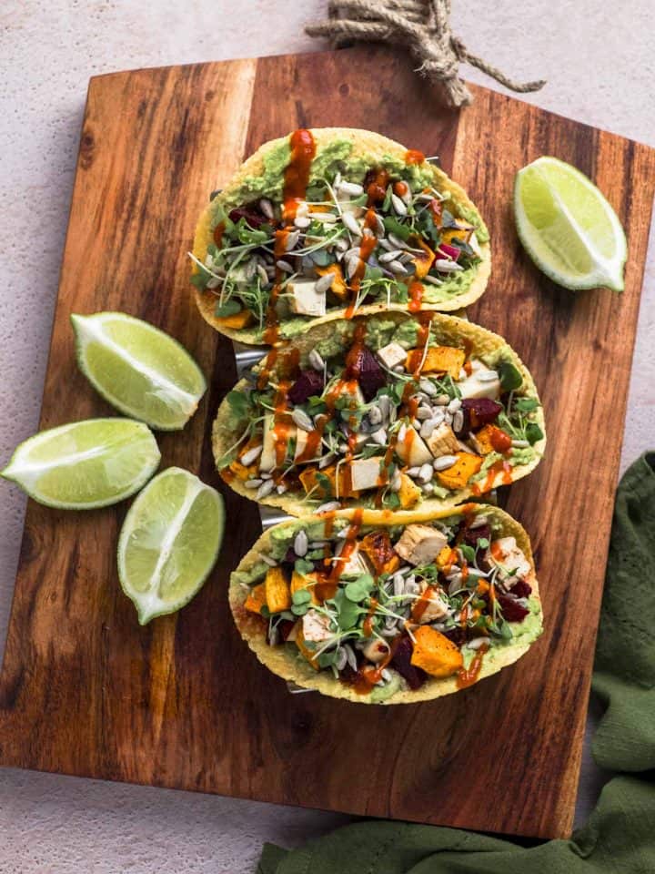 overhead image of a wooden cutting board. 3 corn tortilla tacos are on top of it. The tacos are filled with a mixture of beets, sweet potatoes, apples, tofu and spices.
