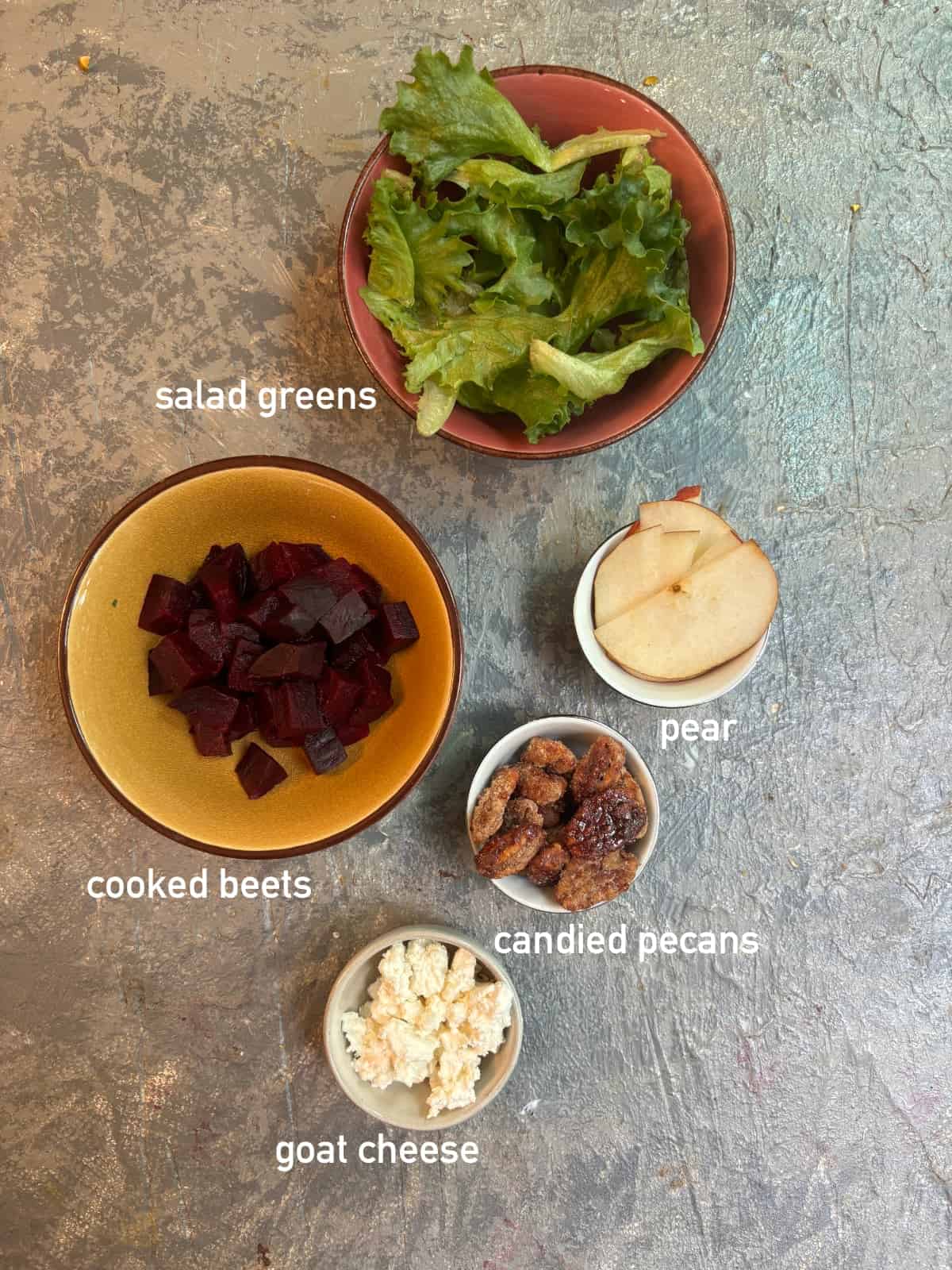 overhead image of the ingredients needed for beet salad. Each ingredient is in it's own small bowl and labeled with white text