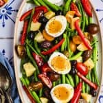 overhead image of an oval serving platter with green bean salad. The salad is a mixture of green beans, grape tomatoes, red bell peppers, olives, potatoes, and hard boiled eggs