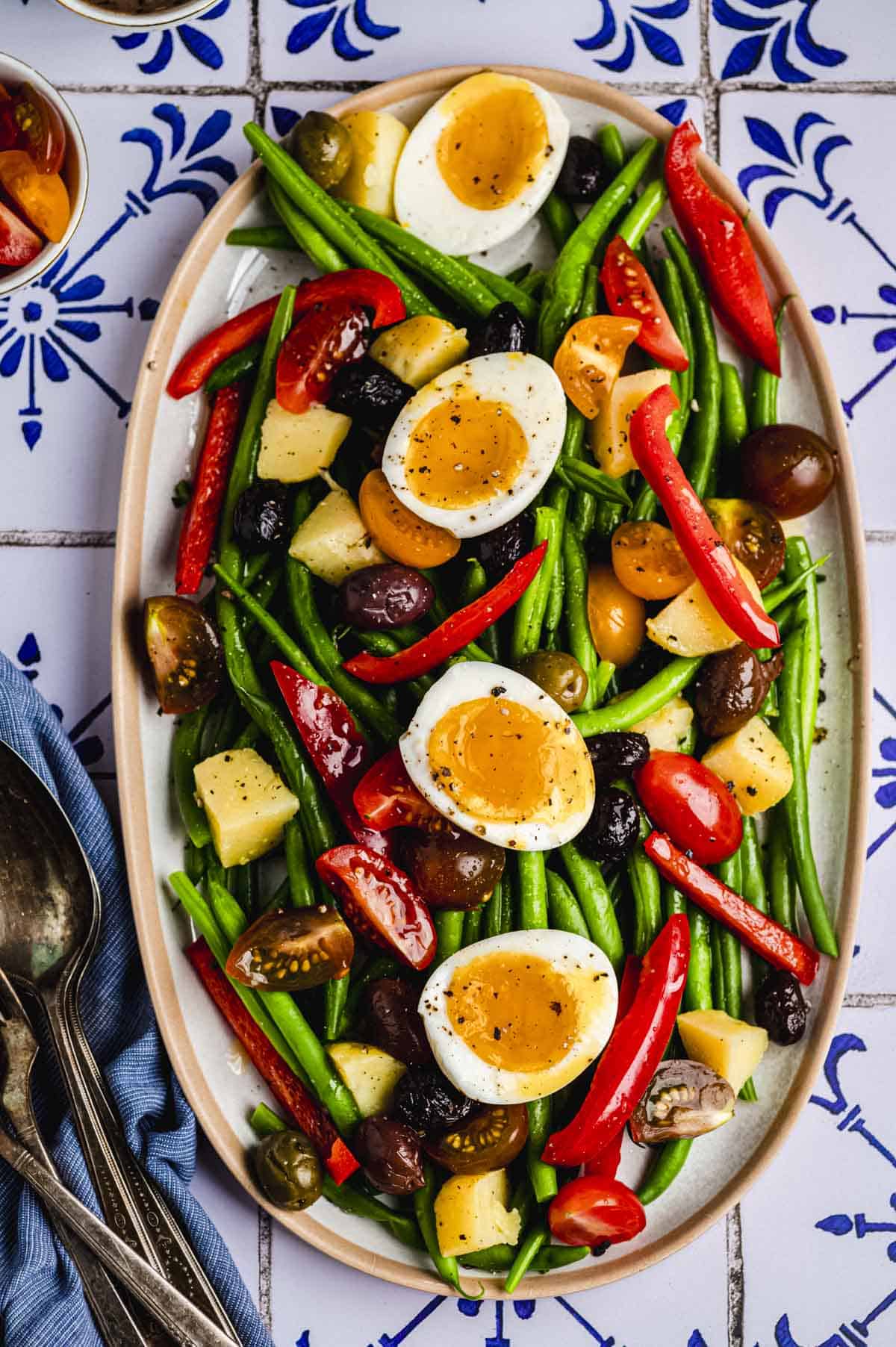 overhead image of an oval serving platter with green bean salad. The salad is a mixture of green beans, grape tomatoes, red bell peppers, olives, potatoes, and hard boiled eggs