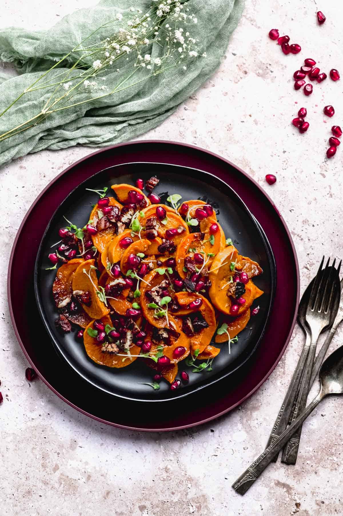 overhead image of a plate of roasted deep orange honeynut squash slices. The squash is topped with poomegranate seeds and micro herbs.