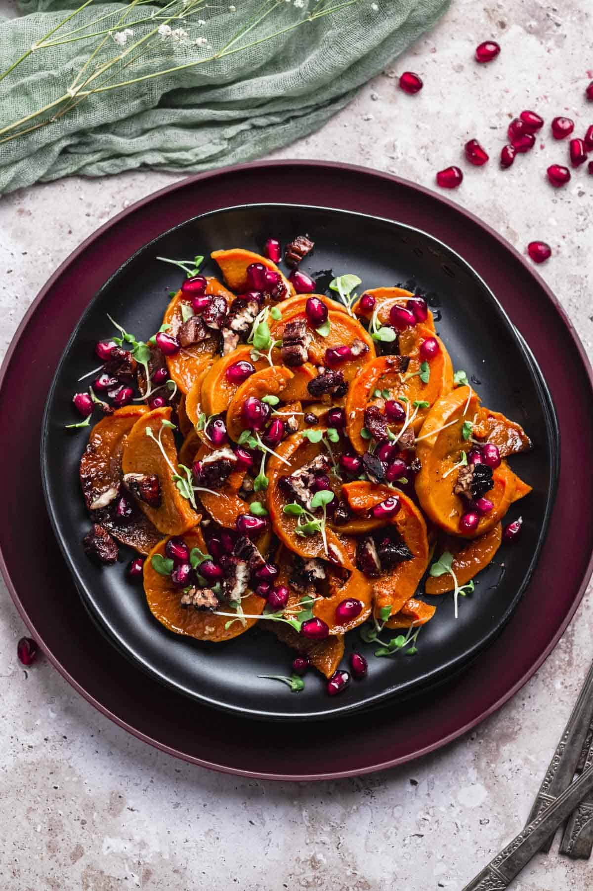 overhead image of a plate of roasted deep orange honeynut squash slices. The squash is topped with poomegranate seeds and micro herbs.