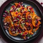 overhead image of a plate of roasted deep orange honeynut squash slices. The squash is topped with poomegranate seeds and micro herbs.