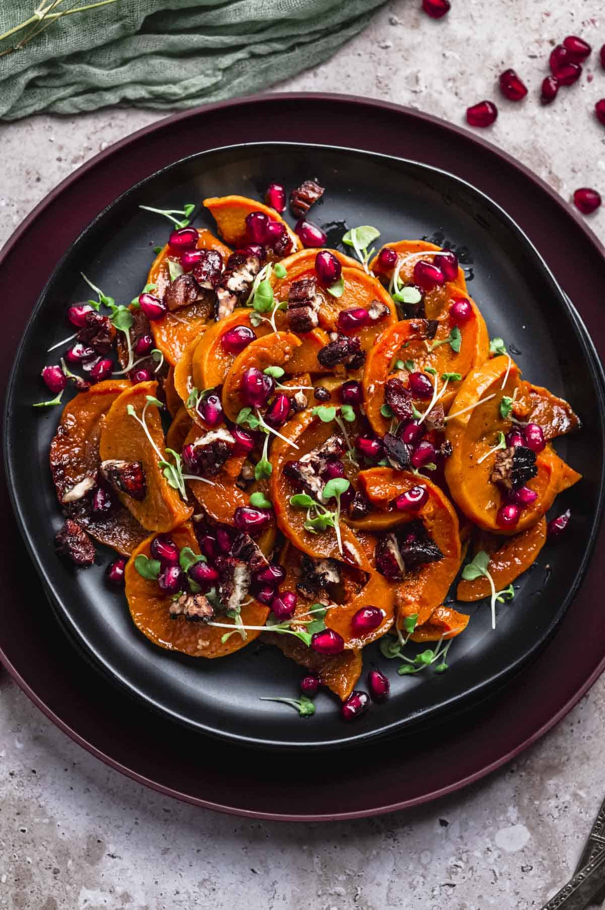 overhead image of a plate of roasted deep orange honeynut squash slices. The squash is topped with poomegranate seeds and micro herbs.