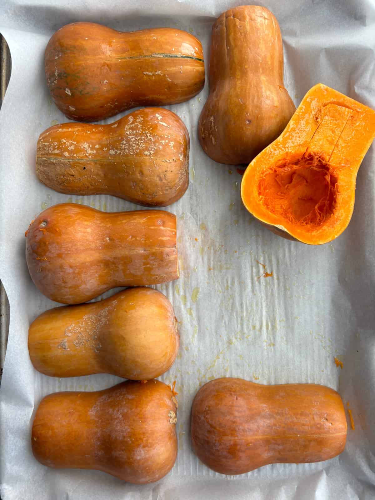 overhead image of 8 halves of honeynut squash laying cut side down on a baking sheet lined with parchment paper. One squash half is cut side up to show that the seeds have been removed.