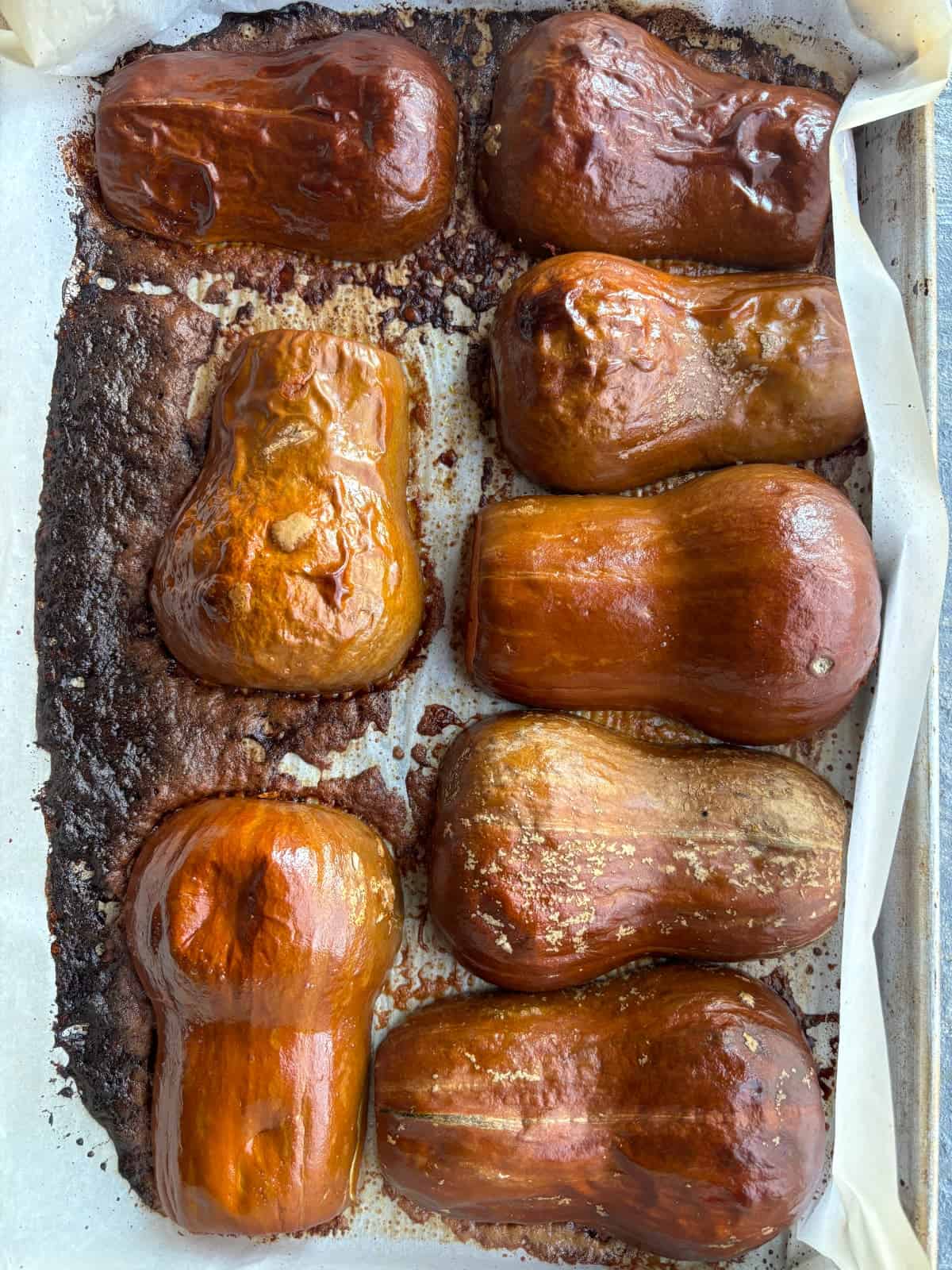 overhead image of 8 halves of honeynut squash laying cut side down on a baking sheet lined with parchment paper. The squash has been roasted, the skins are wrinkled and golden brown and some juice from squash has caramelized on the parchment paper.