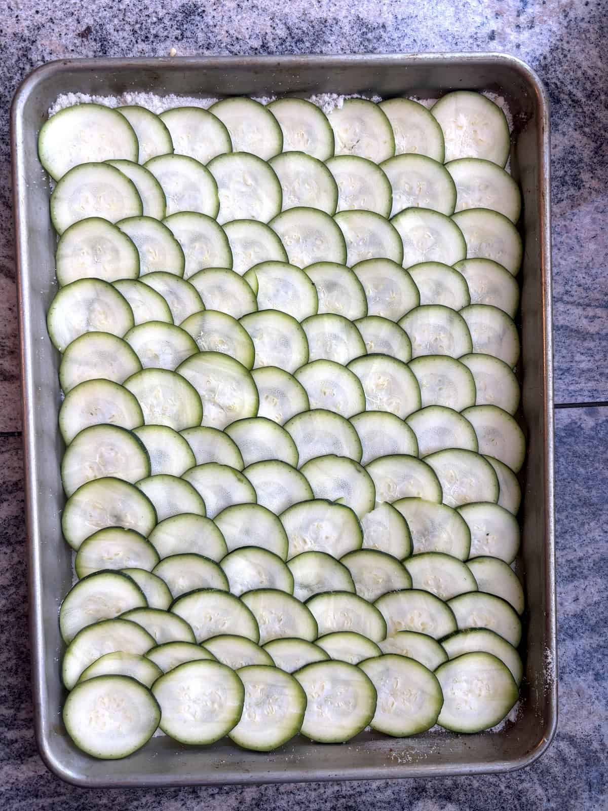 A baking sheet with thinly sliced zucchini that lays of top of layer of grates parmesan cheese