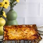 Golden baked lasagna in a white ceramic dish on striped towel with yellow flowers in background.